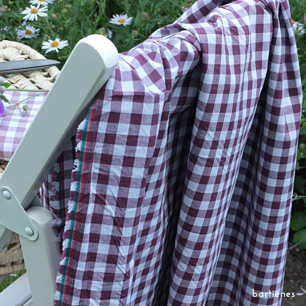 nahaufnahme-drei-karo-stoffe-gestapelt-auf-picknickkorb-im-sommergarten-vor-blumenbeet